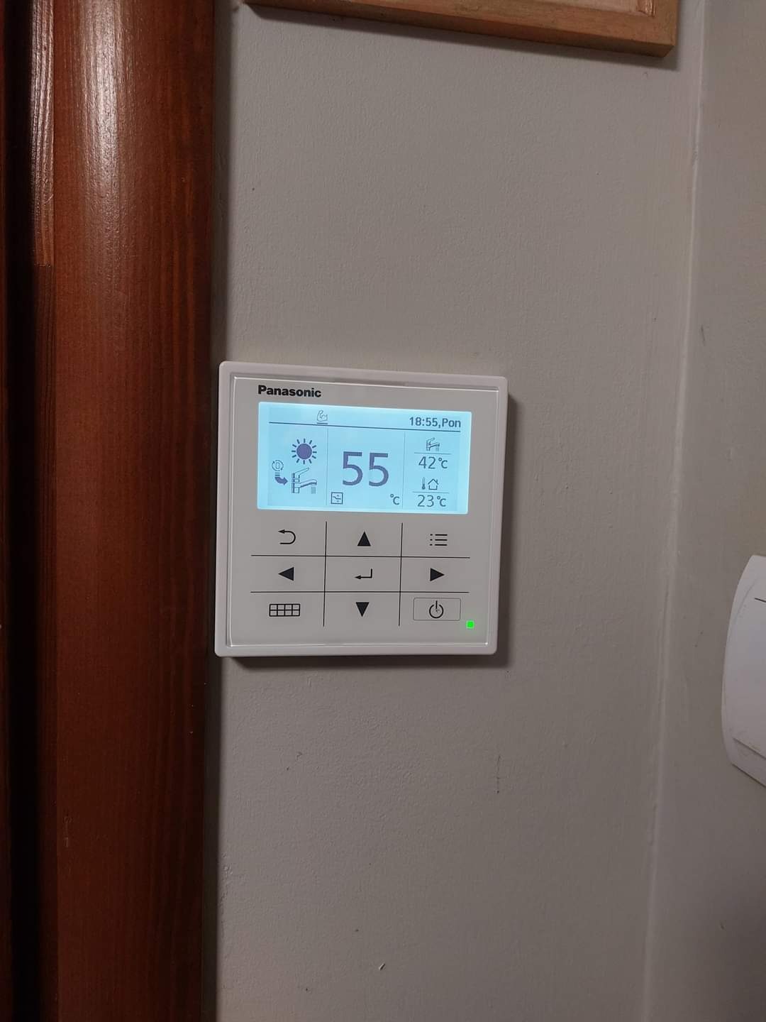 Panel sterowania Panasonic na szarej ścianie, wyświetlający temperaturę z ikonami słońca i kropli wody. Widoczny fragment drewnianej futryny drzwi.