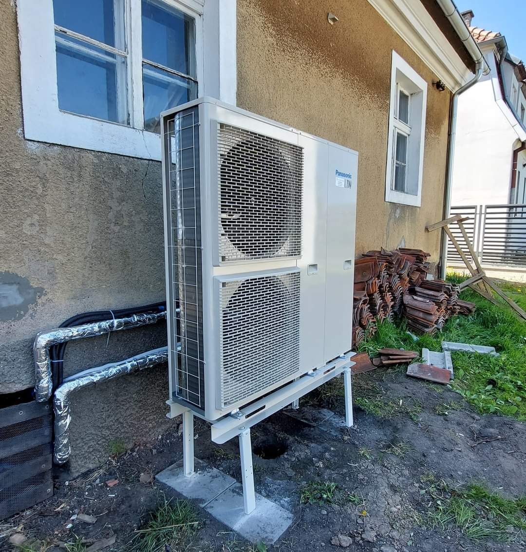 Zewnętrzna jednostka klimatyzacji Panasonic zamontowana na białej, metalowej konstrukcji przy ścianie budynku z widocznymi rurami w izolacji, sterta starych dachówek w tle.