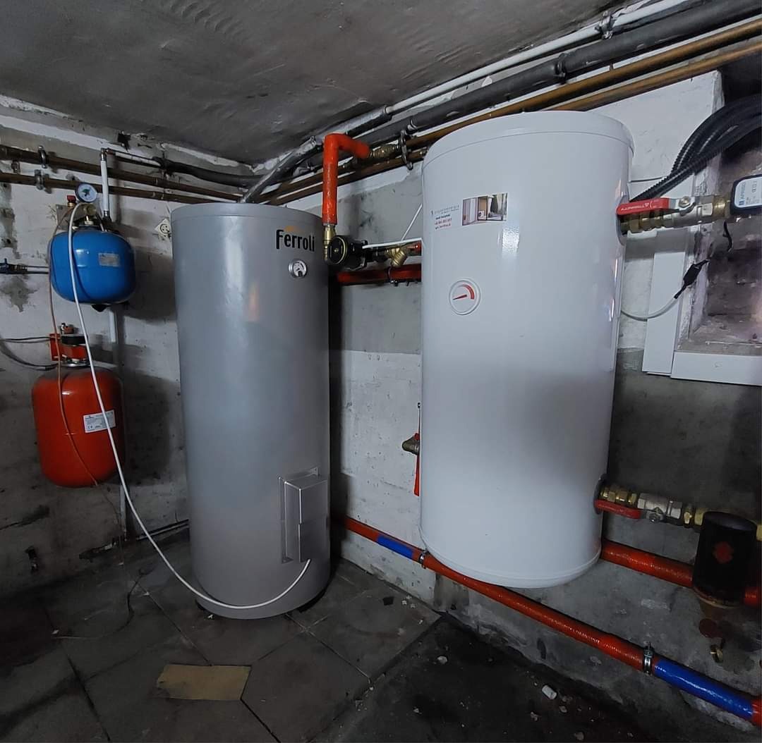 Instalacja dwóch zbiorników: szarego Ferroli i białego, z widocznymi rurami miedzianymi i plastikowymi w kolorze pomarańczowym i niebieskim, zamontowanych w piwnicy z surowym betonowym sufitem.