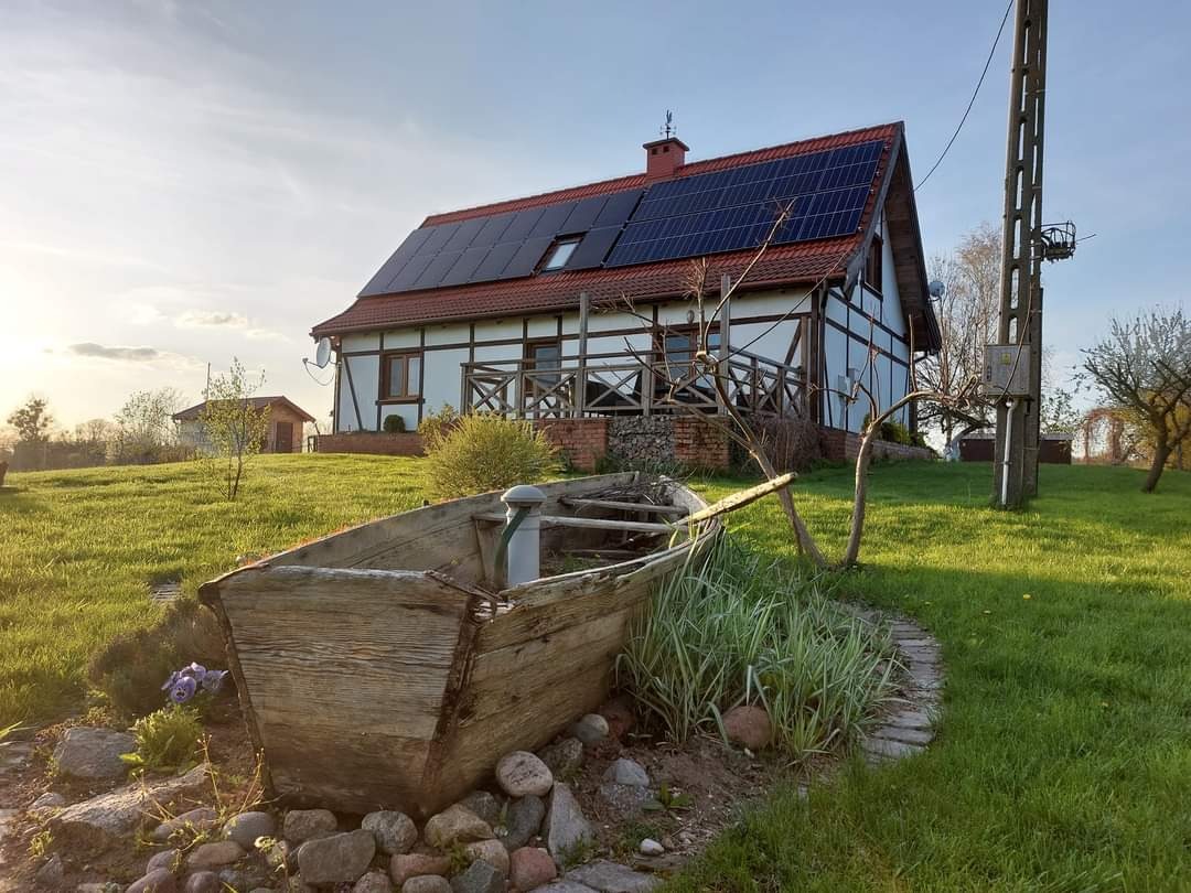 Dom z panelami fotowoltaicznymi na dachu, widok z ogrodu z elementami dekoracyjnymi: stara drewniana łódź i ścieżka z kamieni.