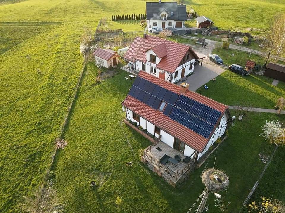 Panoramiczny widok z drona na dom z panelami słonecznymi na dachu, otoczony zielonym trawnikiem, z gniazdem bociana na pierwszym planie i zabudowaniami w tle.