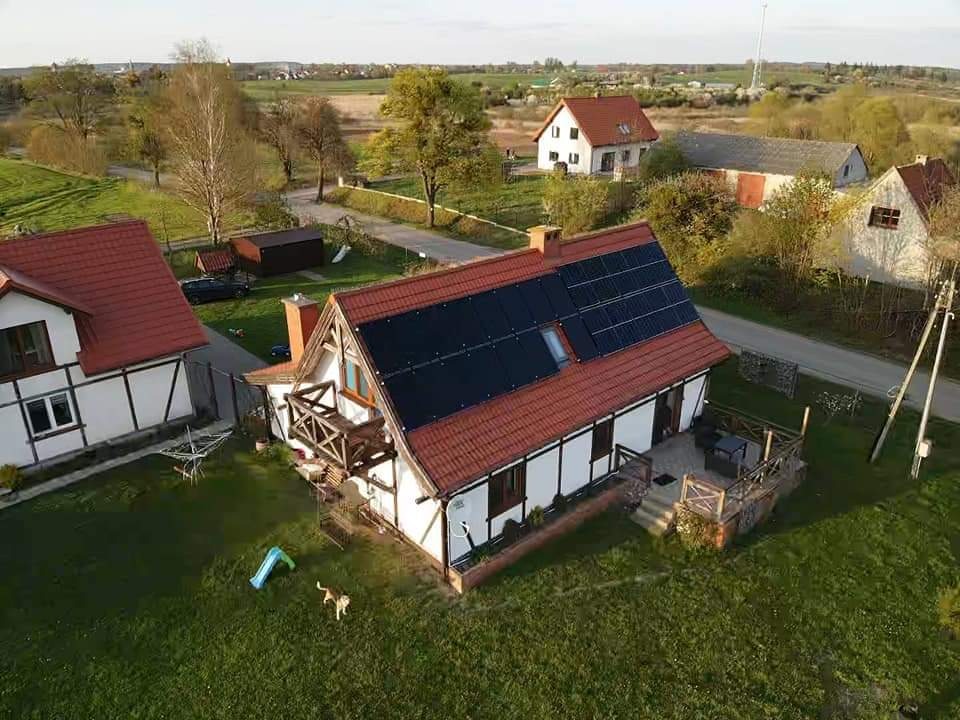 Dom jednorodzinny z czerwoną dachówką i panelami słonecznymi na dachu, widok z lotu ptaka, pies na trawniku, w tle zabudowa wiejska.