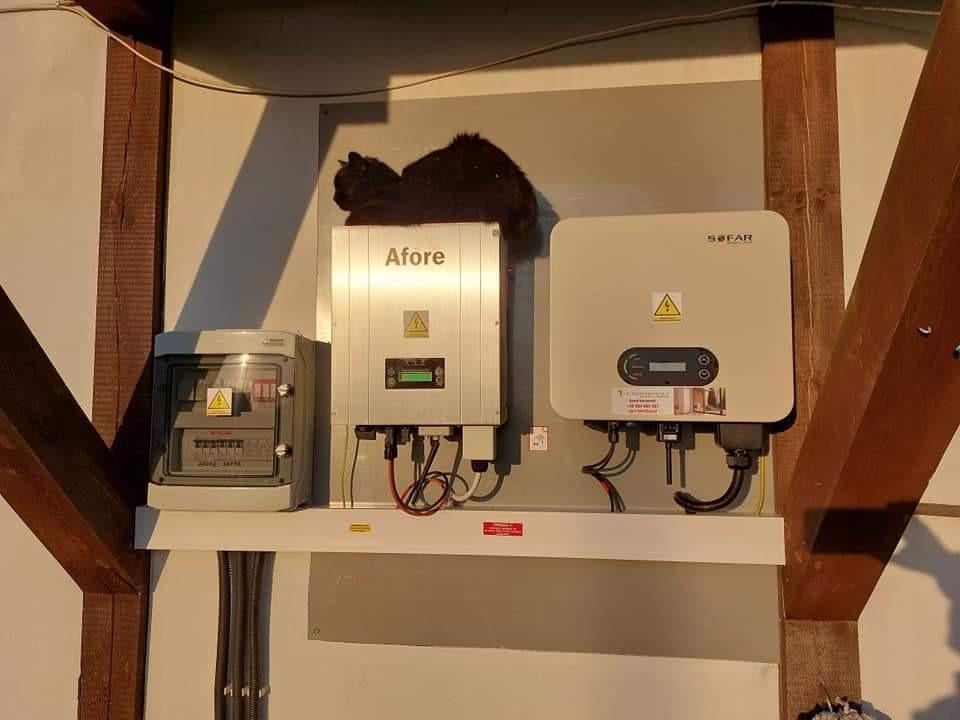 Instalacja fotowoltaiczna z inwerterami Afore i SOFAR oraz czarnym kotem leżącym na górze inwertera Afore, zamontowana na białej ścianie w pobliżu drewnianej belki.