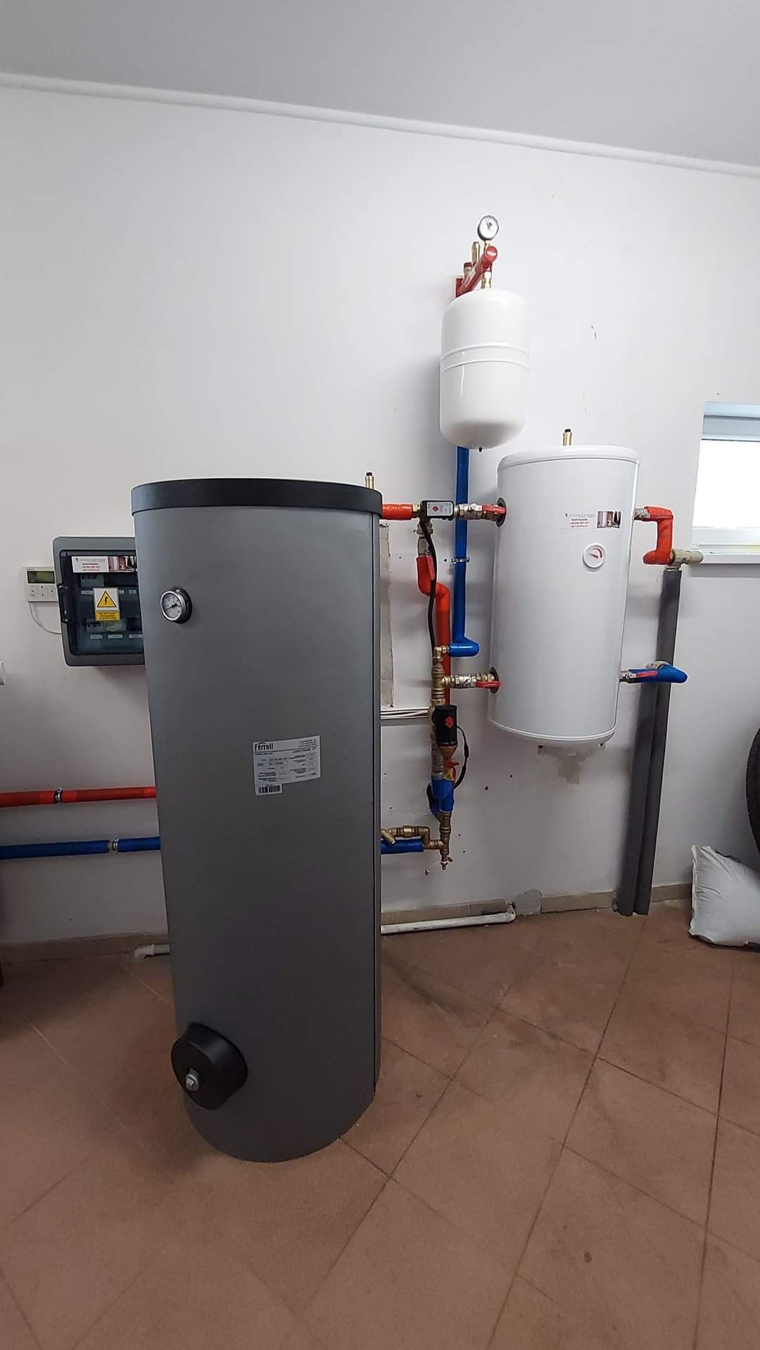 Szary zbiornik akumulacyjny Ferroli z osprzętem hydraulicznym, manometrami i zaworami, zainstalowany w pomieszczeniu z brązową terakotą.