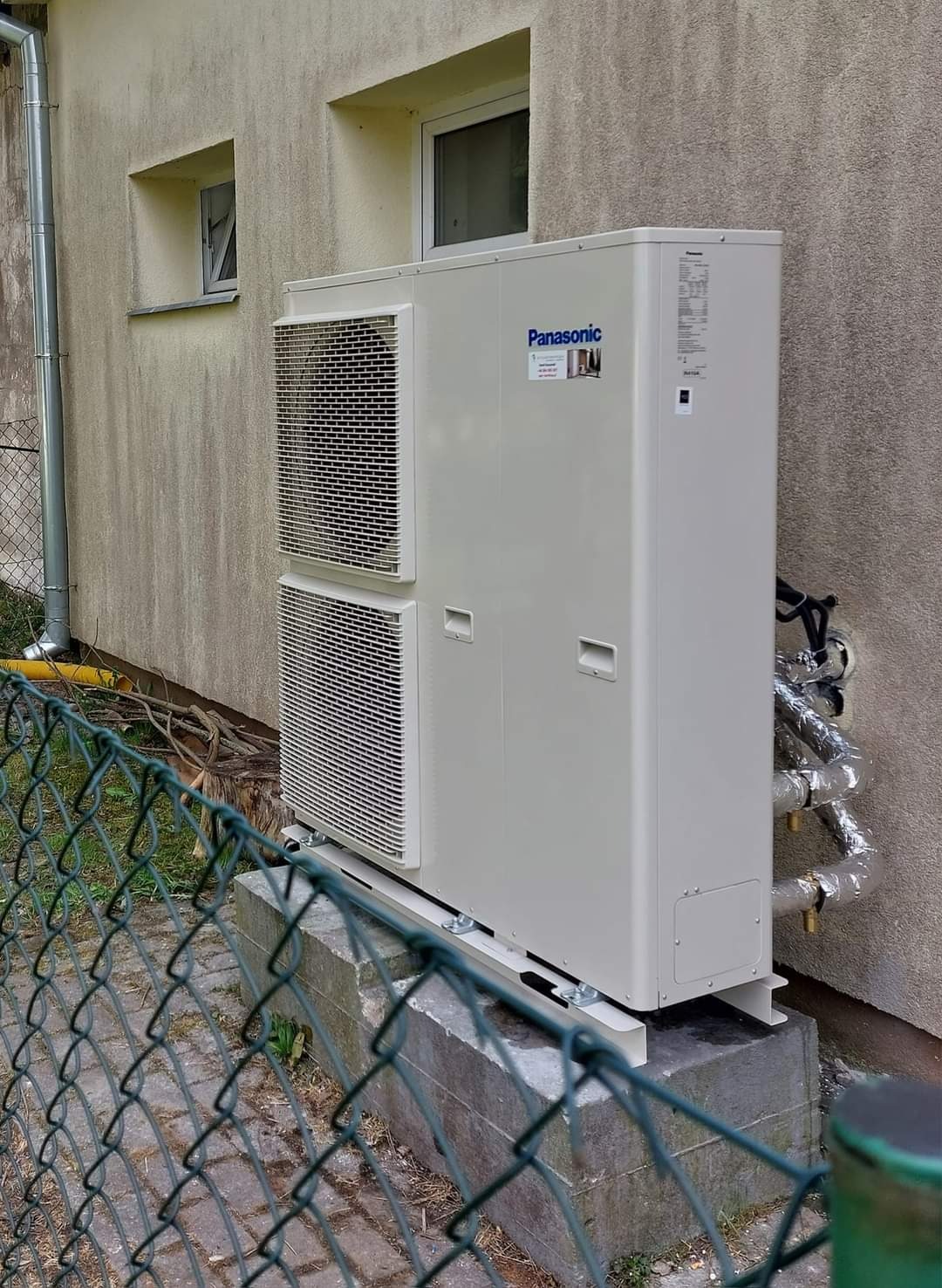 Zewnętrzna jednostka pompy ciepła Panasonic Aquarea zamontowana na betonowym fundamencie przy ścianie budynku z widocznymi rurami w izolacji termicznej.