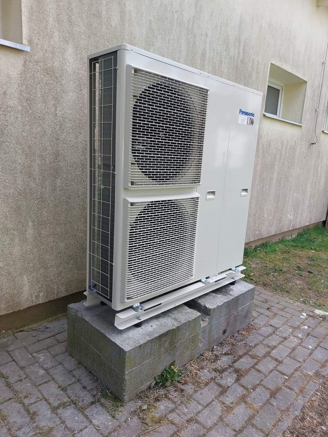 Zewnętrzna jednostka klimatyzacji Panasonic, zamontowana na betonowym fundamencie przy ścianie budynku z widocznym oknem.