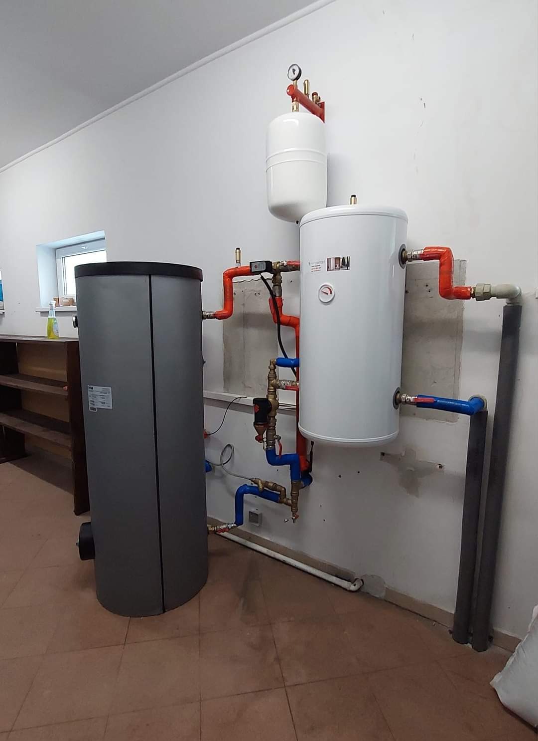 Szara pompa ciepła typu monoblok obok białego zbiornika CWU i mniejszego zbiornika buforowego, połączone rurami miedzianymi i niebieskimi, zamontowane na białej ścianie w pomieszczeniu z brązową...