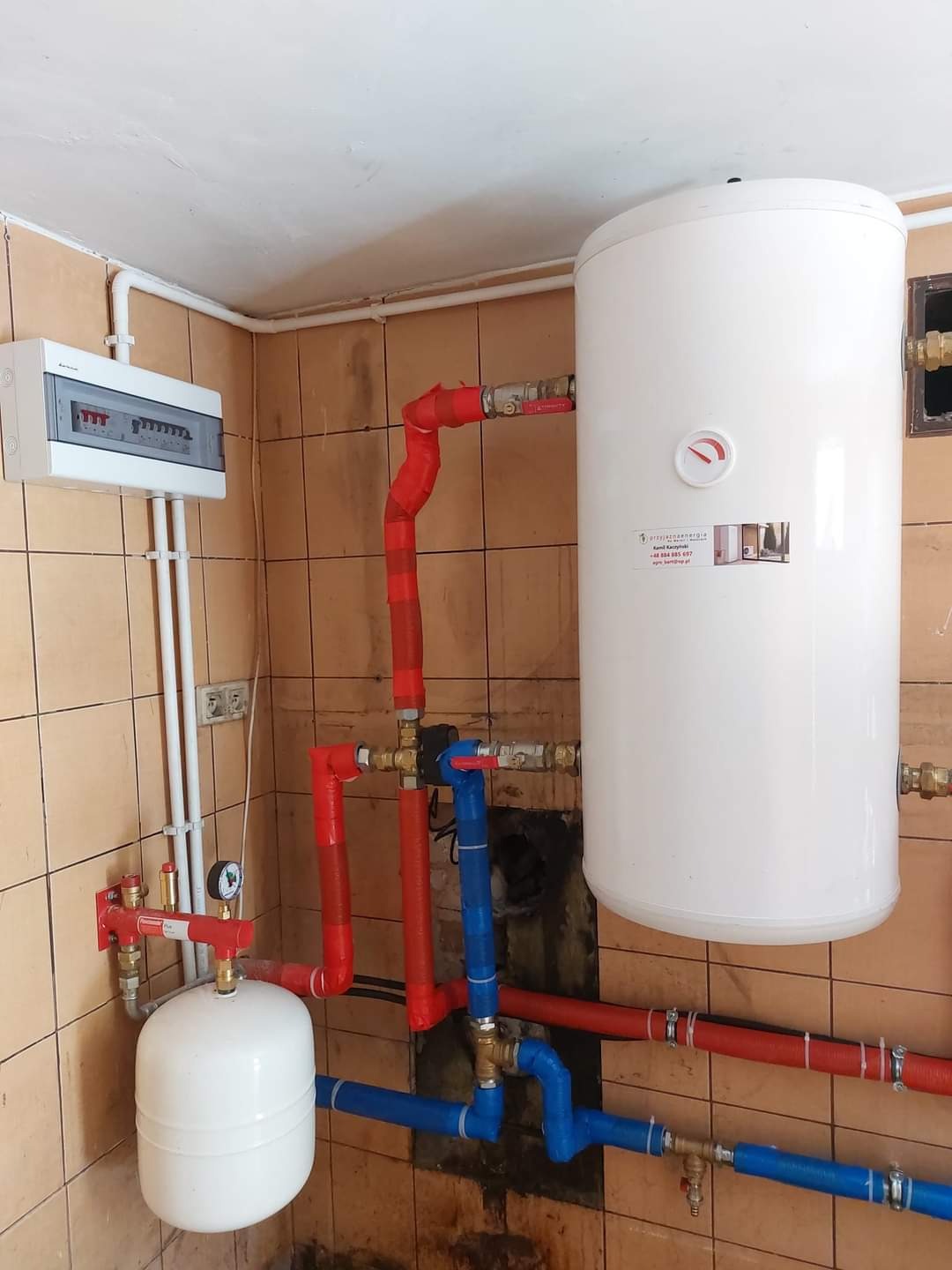 Biały zasobnik ciepłej wody użytkowej z czerwono-niebieską instalacją hydrauliczną w pomieszczeniu z brązowymi płytkami, widoczna skrzynka elektryczna na ścianie.