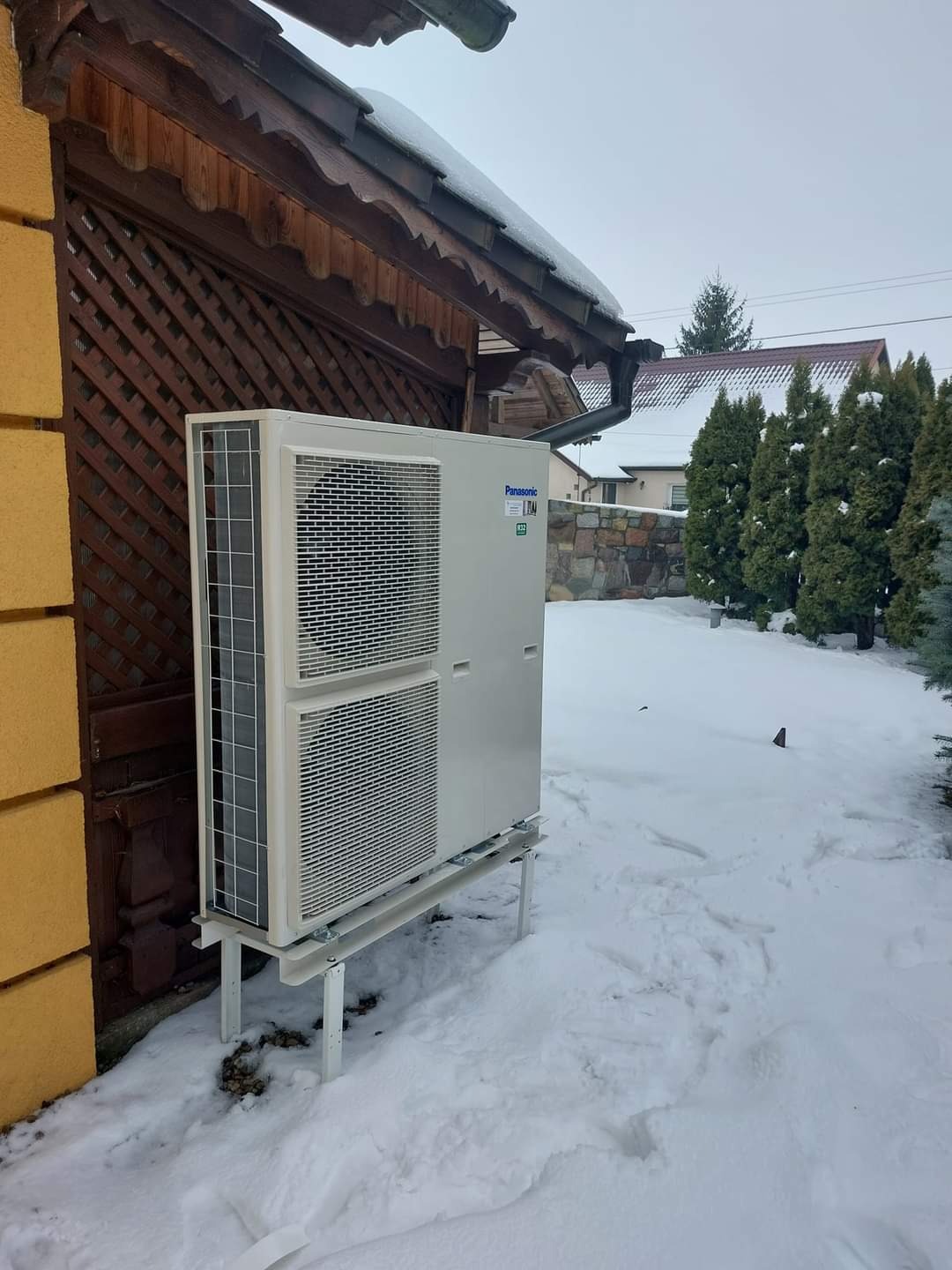 Biała jednostka zewnętrzna klimatyzatora Panasonic, zamontowana na metalowym stelażu przy drewnianej altanie w zimowym krajobrazie, śnieg na dachu i ziemi.