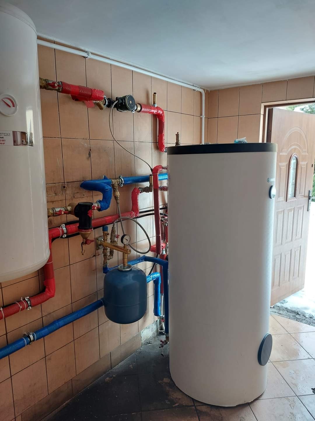 Instalacja bufora i zbiornika wody użytkowej z licznymi rurami w kolorach czerwonym i niebieskim, widoczne w pomieszczeniu w Pasym, z drzwiami prowadzącymi na zewnątrz.
