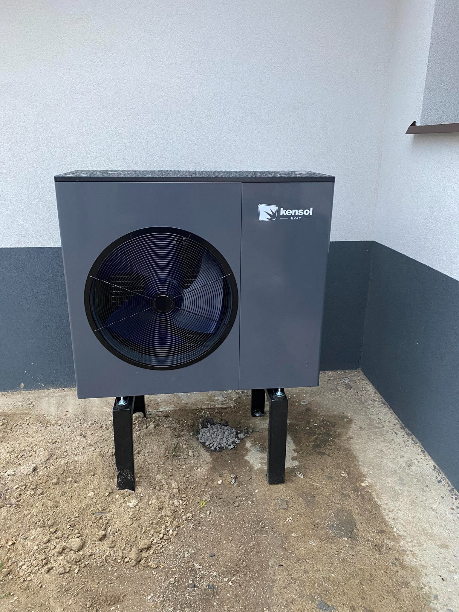 Szara jednostka zewnętrzna z wentylatorem marki Kensol HVAC, umieszczona na czarnych wspornikach, stoi na betonowej powierzchni przy ścianie budynku.