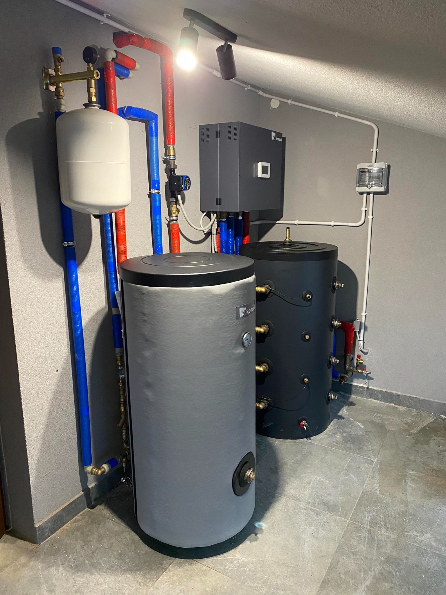 Dwa zbiorniki buforowe, szary i czarny, z podłączonymi rurami w kolorze czerwonym i niebieskim, zamontowane w rogu pomieszczenia z szarą podłogą i ścianami. Widoczny również biały zbiornik...