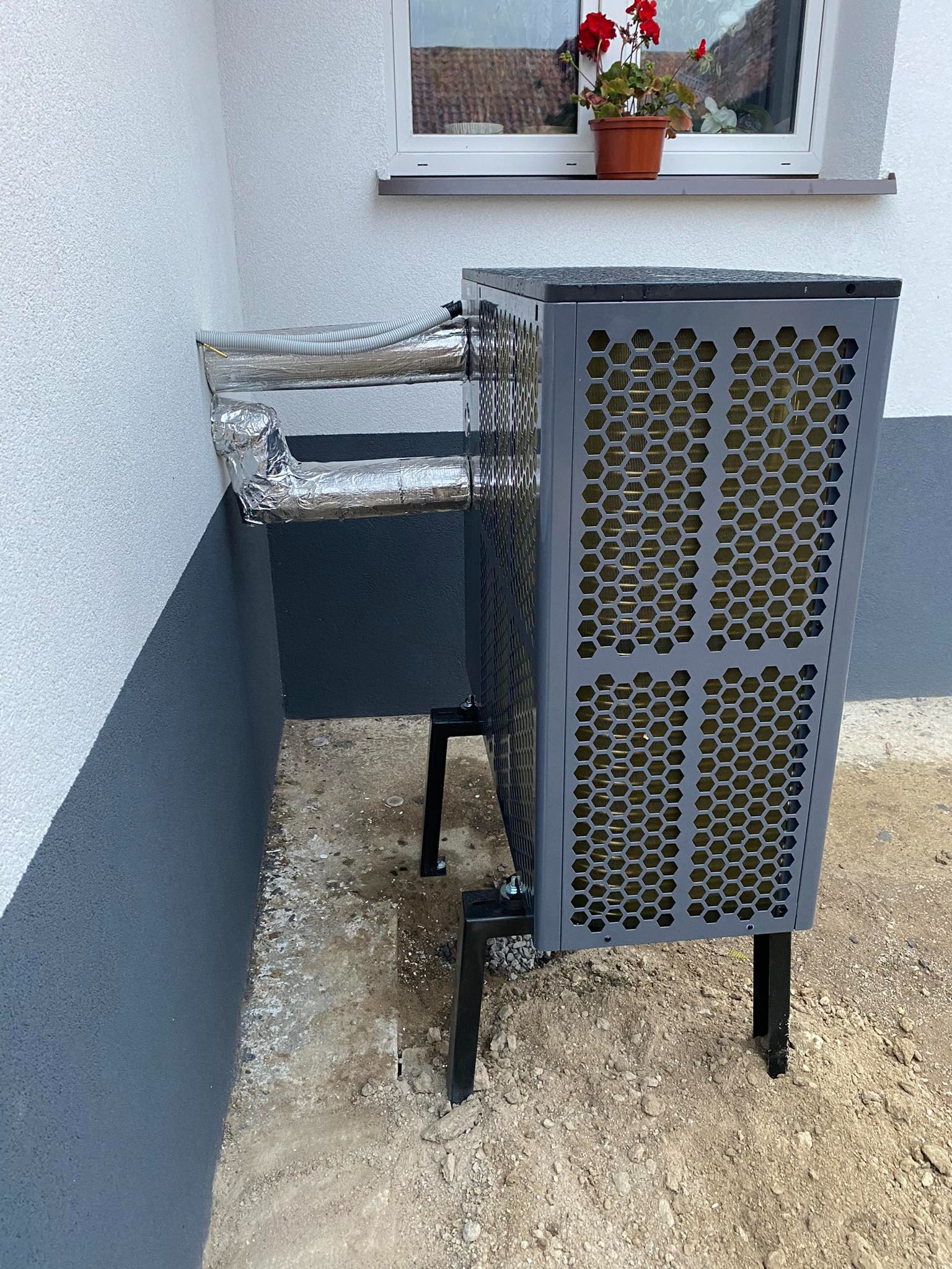 Szara pompa ciepła na czarnych nóżkach, zainstalowana przy ścianie budynku z widocznymi izolowanymi rurami, z doniczką z czerwonymi kwiatami na parapecie okna w tle.