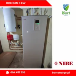 Biała pompa ciepła Nibe zainstalowana w pomieszczeniu z widocznymi instalacjami hydraulicznymi i zbiornikiem buforowym, z napisem 'BOCHLIN 8 KW' w górnej części zdjęcia.