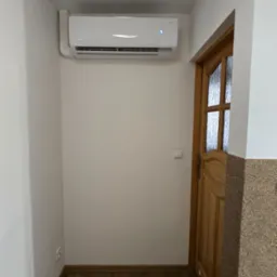 Biały klimatyzator ścienny zamontowany w korytarzu nad drewnianymi drzwiami. Widoczny wyświetlacz temperatury. Jasne ściany i drewnopodobna podłoga.