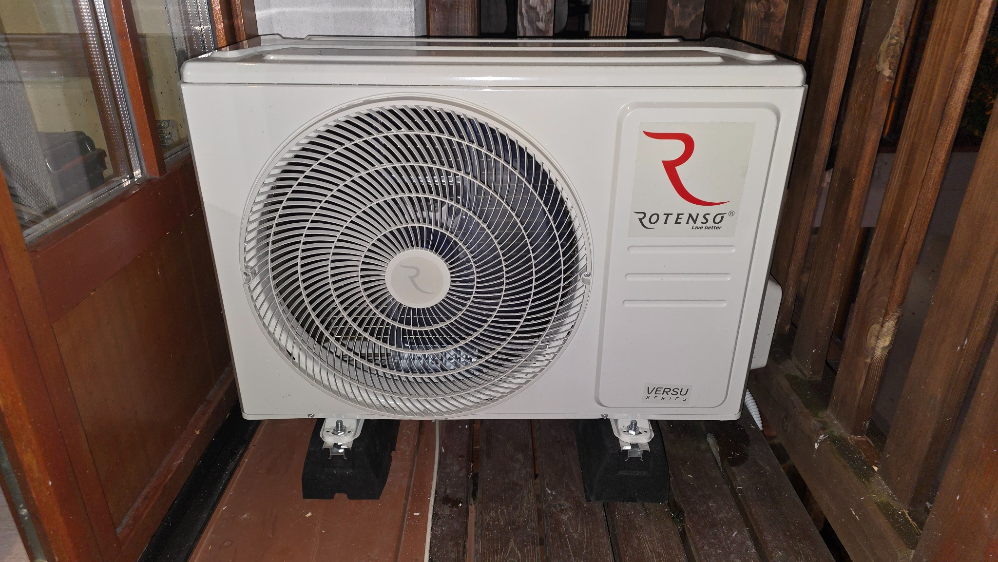 Biała, zewnętrzna jednostka klimatyzacji Rotenso Versu Series, zamontowana na drewnianym balkonie, widoczna z bliska, z elementami drewnianej balustrady i drzwi.