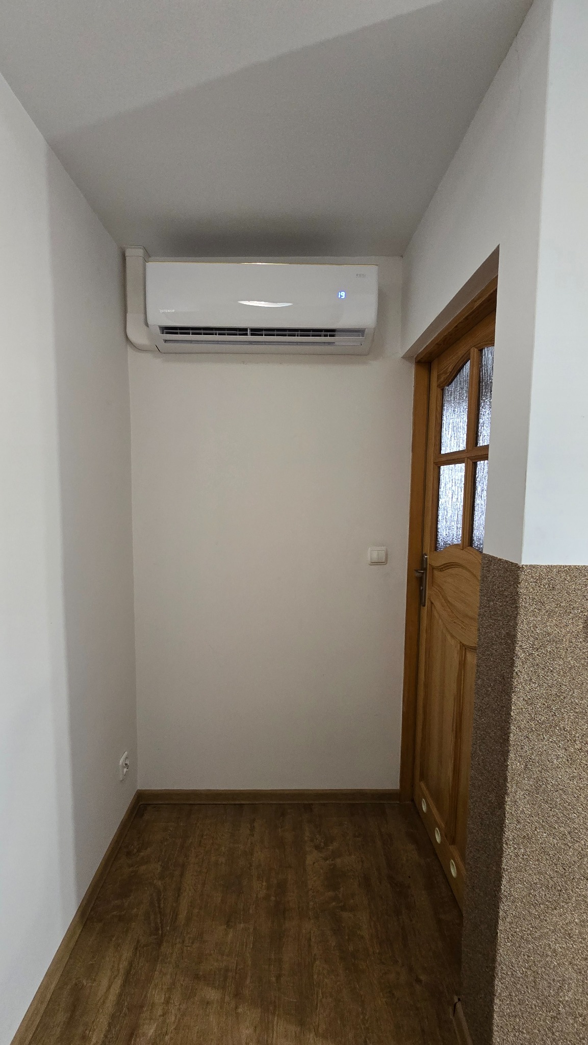 Biały klimatyzator ścienny zamontowany w korytarzu nad drewnianymi drzwiami. Widoczny wyświetlacz temperatury. Jasne ściany i drewnopodobna podłoga.