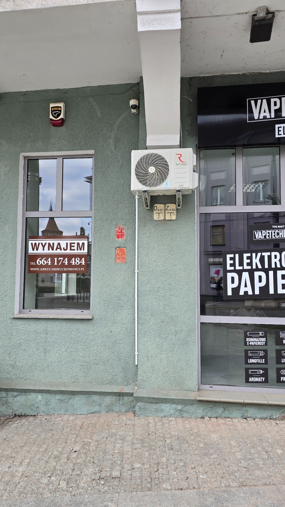 Klimatyzator Rotenso zamontowany na elewacji budynku obok okna z ogłoszeniem 'Wynajem' i witryny sklepu z e-papierosami. Widoczne numery adresowe.