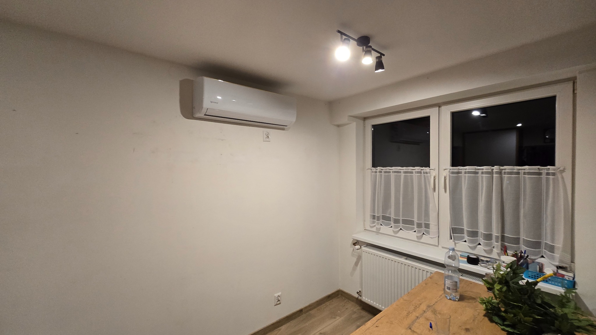 Biały klimatyzator ścienny zamontowany w pokoju z oknem, podświetlony przez lampę sufitową. Widoczny fragment drewnianego blatu i grzejnika pod oknem.