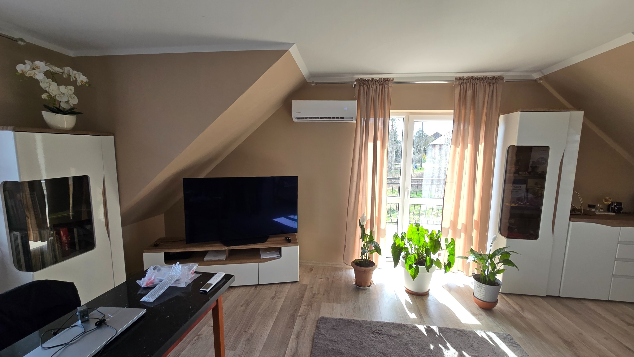 Klimatyzator ścienny w jasnym pokoju na poddaszu, widoczny nad telewizorem. Okno z jasnymi zasłonami w tle. Rośliny doniczkowe dopełniają wystrój.