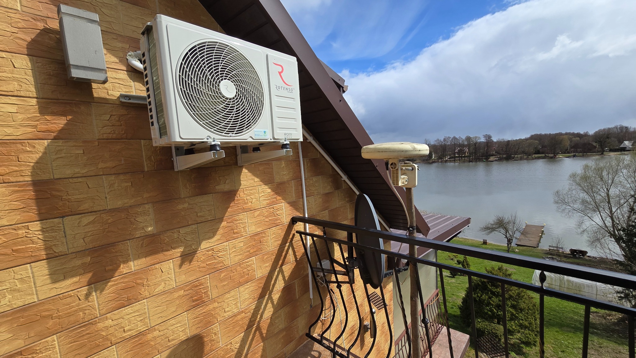 Biała jednostka zewnętrzna klimatyzacji Rotenso zamontowana na ceglanej ścianie budynku z widokiem na jezioro z balkonu. Widoczne elementy instalacji i uchwyty montażowe.