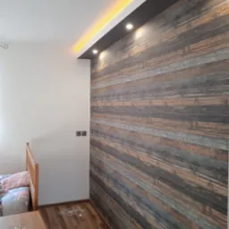 Pokój z drewnopodobną tapetą na jednej ścianie, podświetlonym sufitem i fragmentem łóżka dziecięcego.