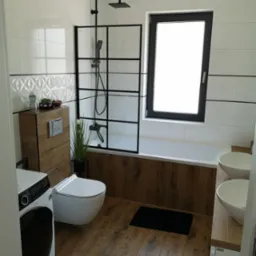 Nowoczesna łazienka z drewnopodobnymi płytkami podłogowymi i akcentami drewna na zabudowie toalety i wannie, białe płytki ścienne z geometrycznym wzorem, czarna rama kabiny prysznicowej i okna...