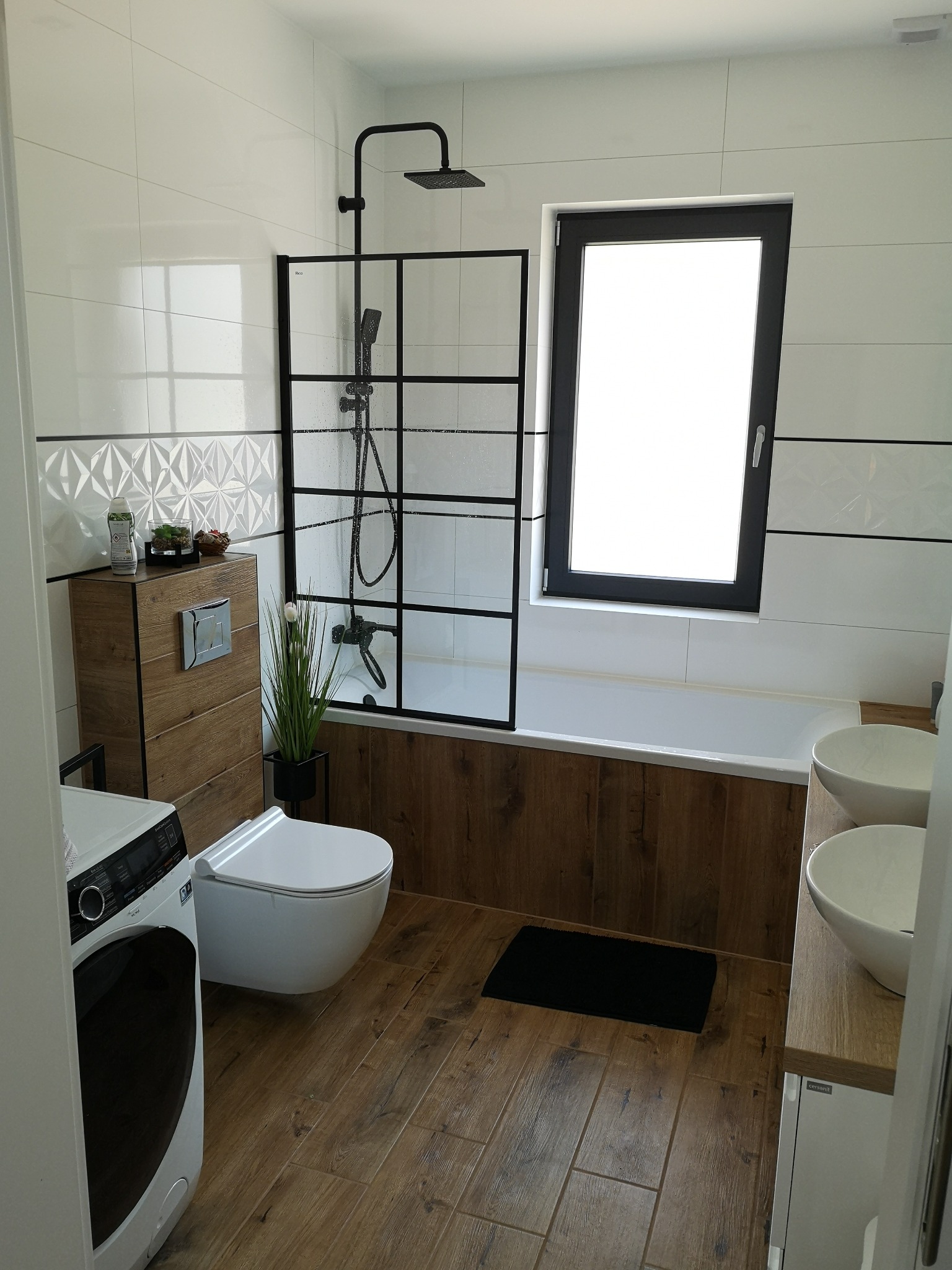Nowoczesna łazienka z drewnopodobnymi płytkami podłogowymi i akcentami drewna na zabudowie toalety i wannie, białe płytki ścienne z geometrycznym wzorem, czarna rama kabiny prysznicowej i okna...