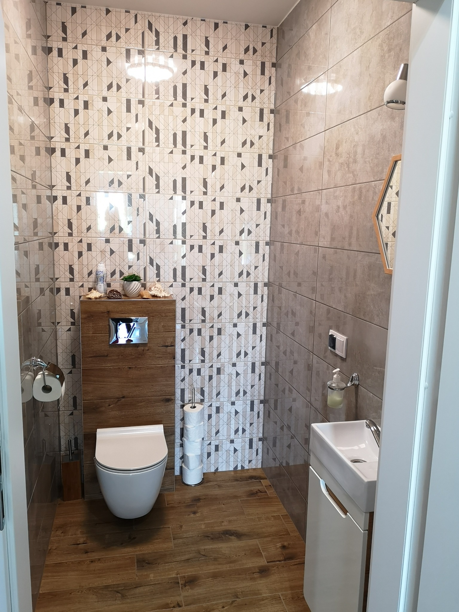 Nowoczesna łazienka z geometrycznymi płytkami ściennymi w odcieniach szarości i beżu, drewnopodobną podłogą, wiszącą toaletą z drewnianą obudową spłuczki i dekoracjami, oraz małą umywalką z szafką.