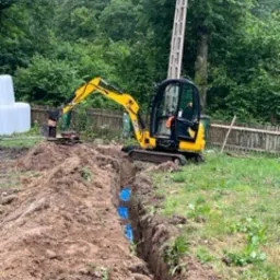 Żółta mini koparka pracująca w wąskim wykopie, widoczne niebieskie rury w ziemi, w tle zieleń drzew i słup energetyczny.