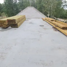 Przygotowane drewniane belki na betonowym stropie domu w budowie, z widocznym szczytem z bloczków betonowych w tle.