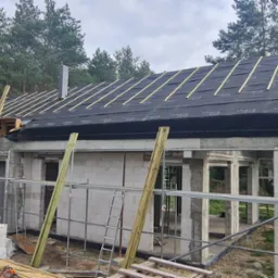 Budowa domu z betonu komórkowego w trakcie wznoszenia dachu, widoczna konstrukcja więźby pokryta membraną dachową i pracownik na rusztowaniu, w tle las.