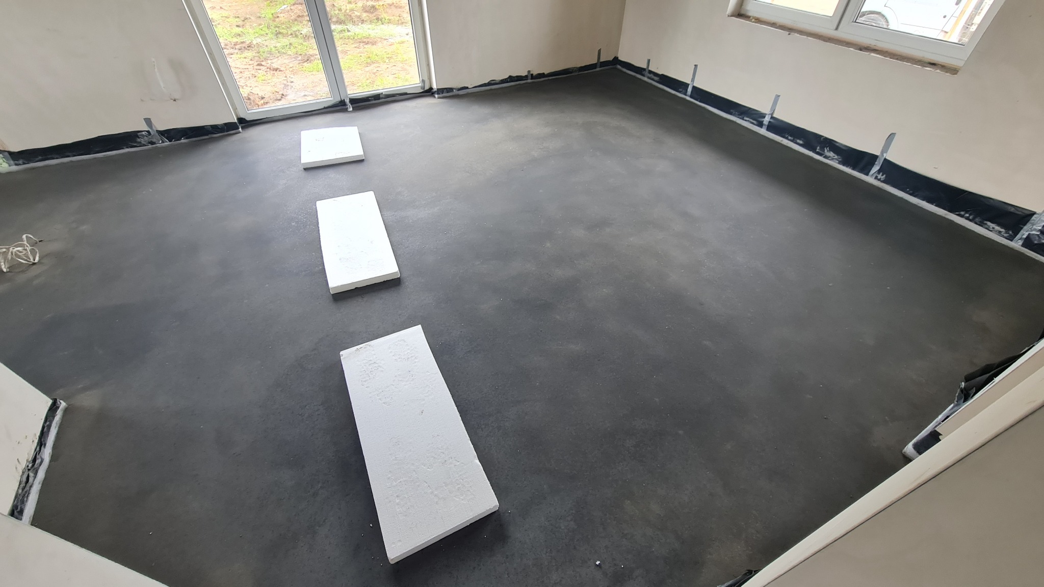 Wylewka podłogowa w surowym wnętrzu z widocznymi płytami styropianowymi, folią izolacyjną przy ścianach i fragmentem okna.