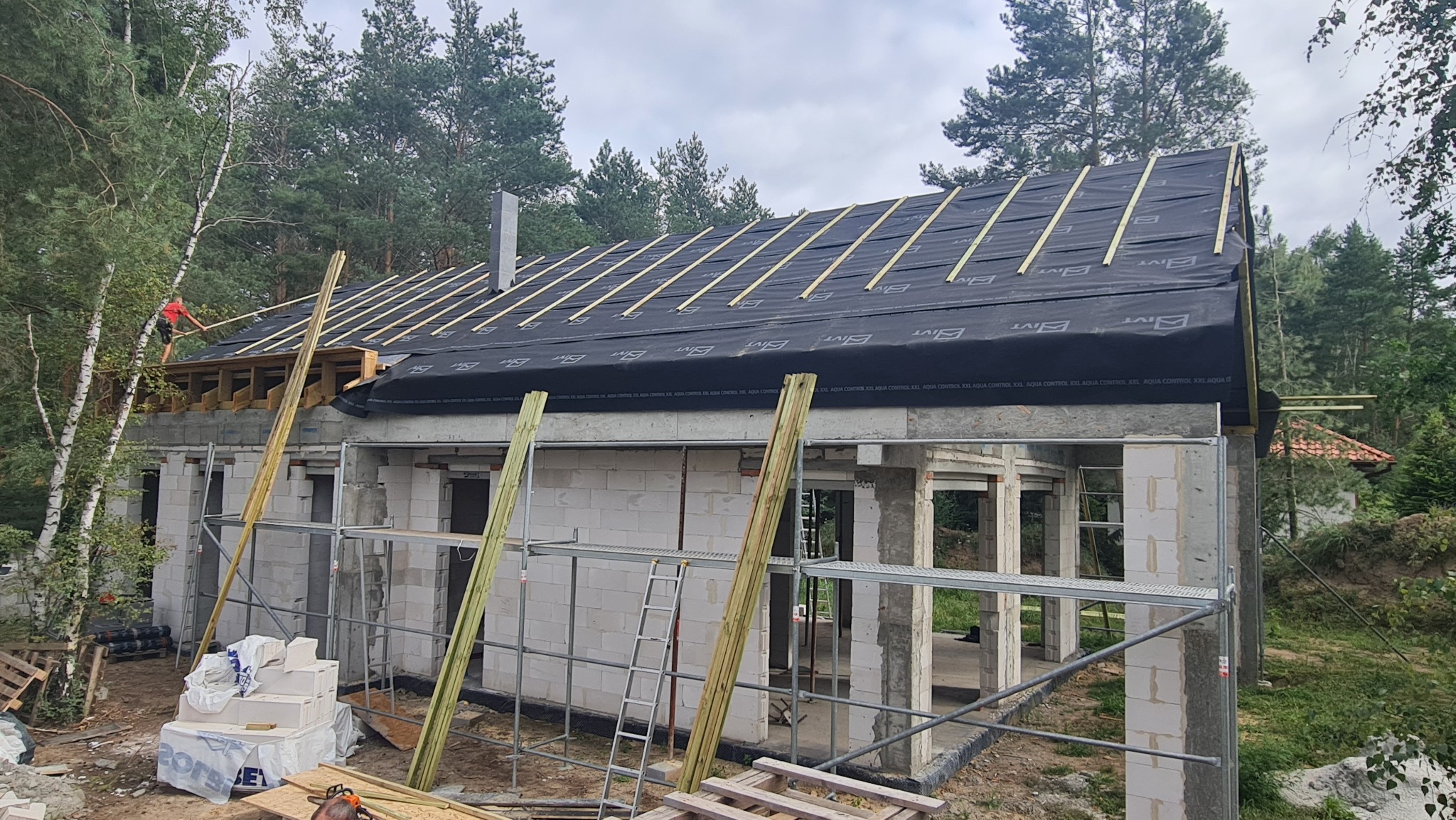 Budowa domu z betonu komórkowego w trakcie wznoszenia dachu, widoczna konstrukcja więźby pokryta membraną dachową i pracownik na rusztowaniu, w tle las.