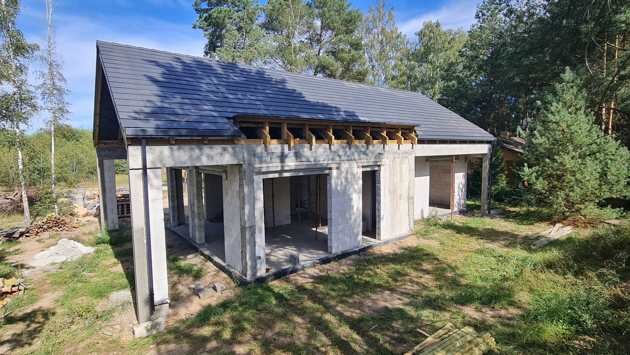 Budowa domu w Poznaniu na etapie wznoszenia ścian i układania ciemnej dachówki, widoczne drewniane elementy konstrukcji dachu i betonowe słupy.