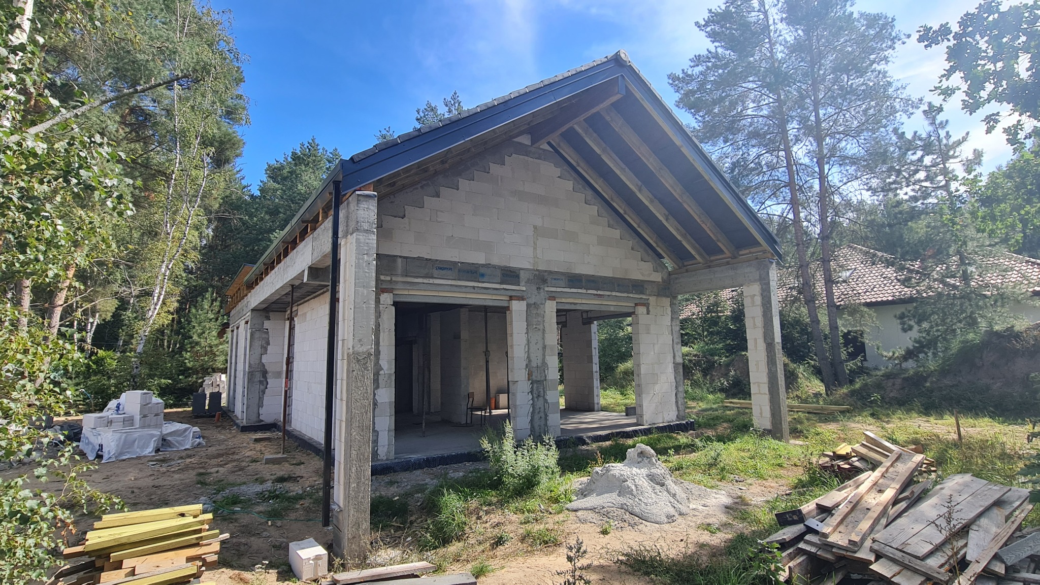 Surowy stan domu murowanego z betonu komórkowego z widoczną konstrukcją dachu, belkami i podciągami, na tle lasu i błękitnego nieba.