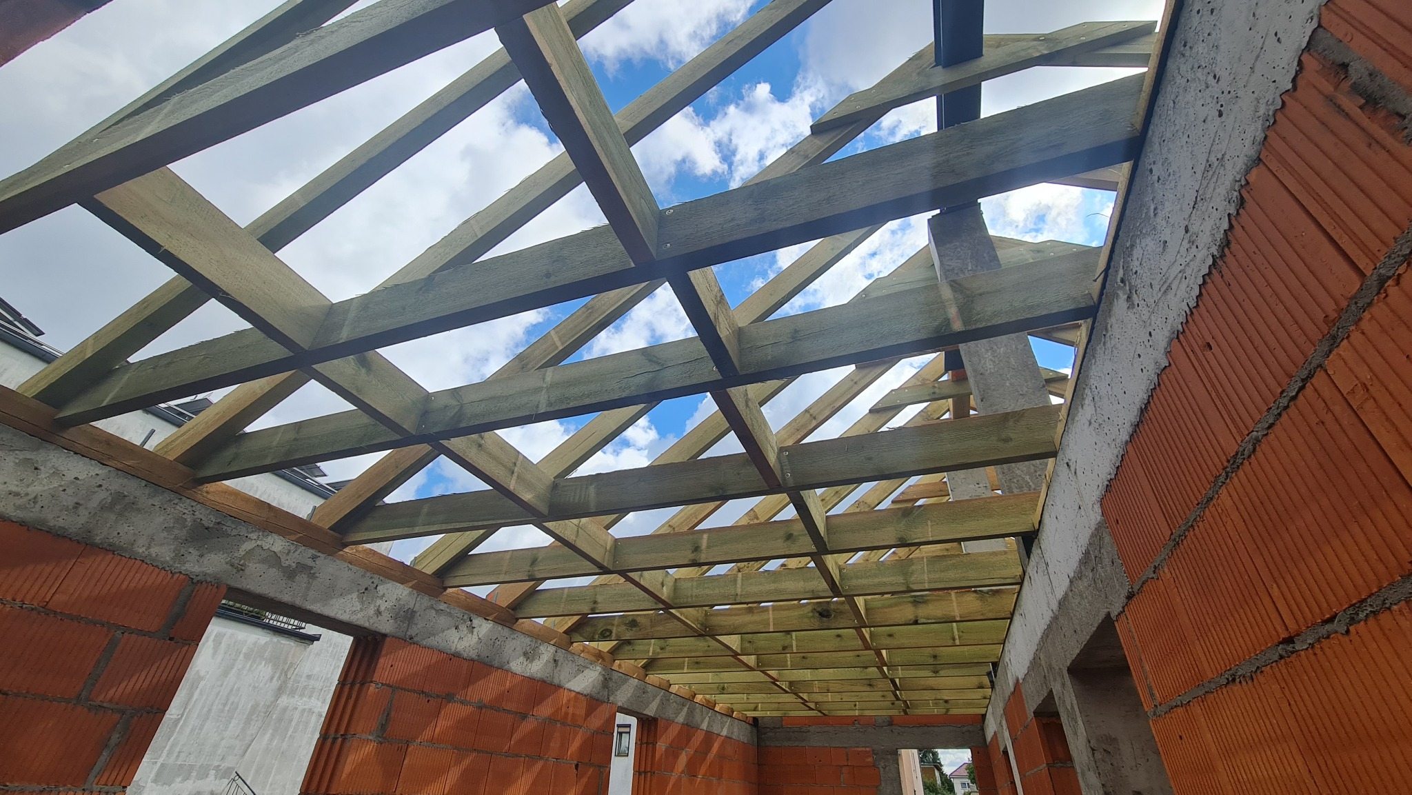 Konstrukcja drewniana dachu w surowym stanie, widok od wewnątrz, z widocznymi ceglanymi ścianami i fragmentem nieba w tle.