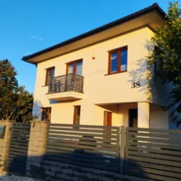 Dwupiętrowy dom z beżową elewacją, balkonem z metalową balustradą, brązowymi oknami i numerem domu 38, widziany zza szarego, poziomego ogrodzenia z kamiennymi słupkami.
