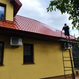 Pracownik na rusztowaniu czyści czerwoną, metalową dachówkę myjką ciśnieniową; żółta elewacja budynku z oknami i klimatyzatorem w tle.