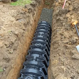 Długi rów wykopany w ziemi z ułożonymi modułowymi tunelami rozsączającymi, podsypany żwirem. Widoczny fragment koparki i poziomica.