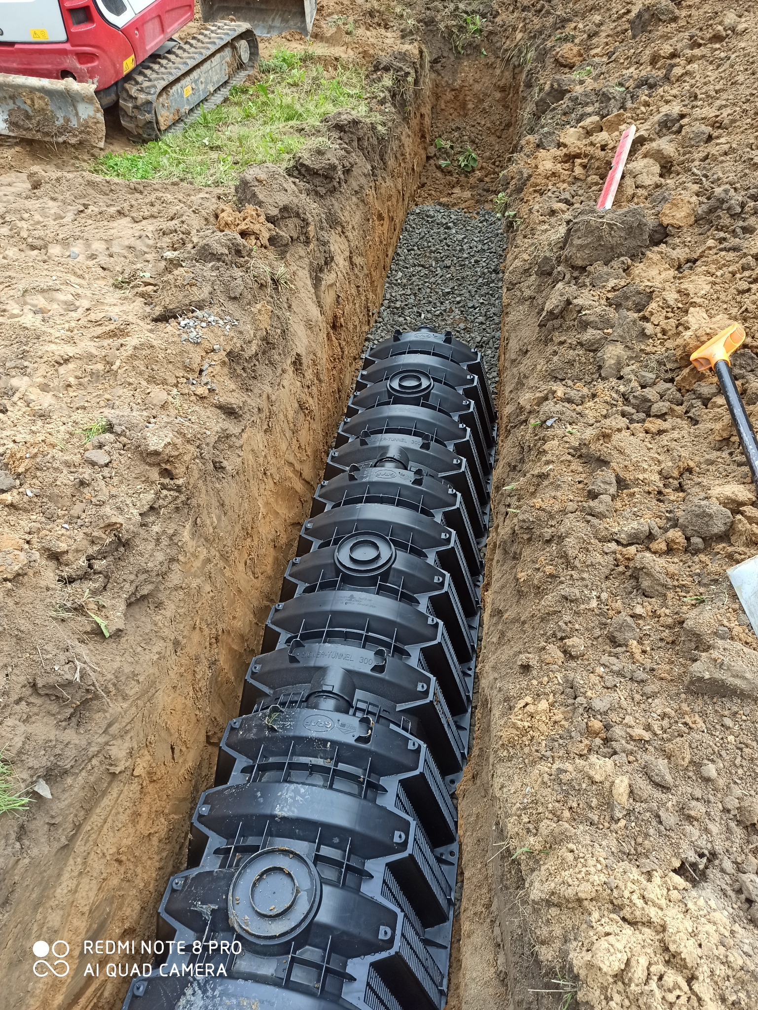 Długi rów wykopany w ziemi z ułożonymi modułowymi tunelami rozsączającymi, podsypany żwirem. Widoczny fragment koparki i poziomica.