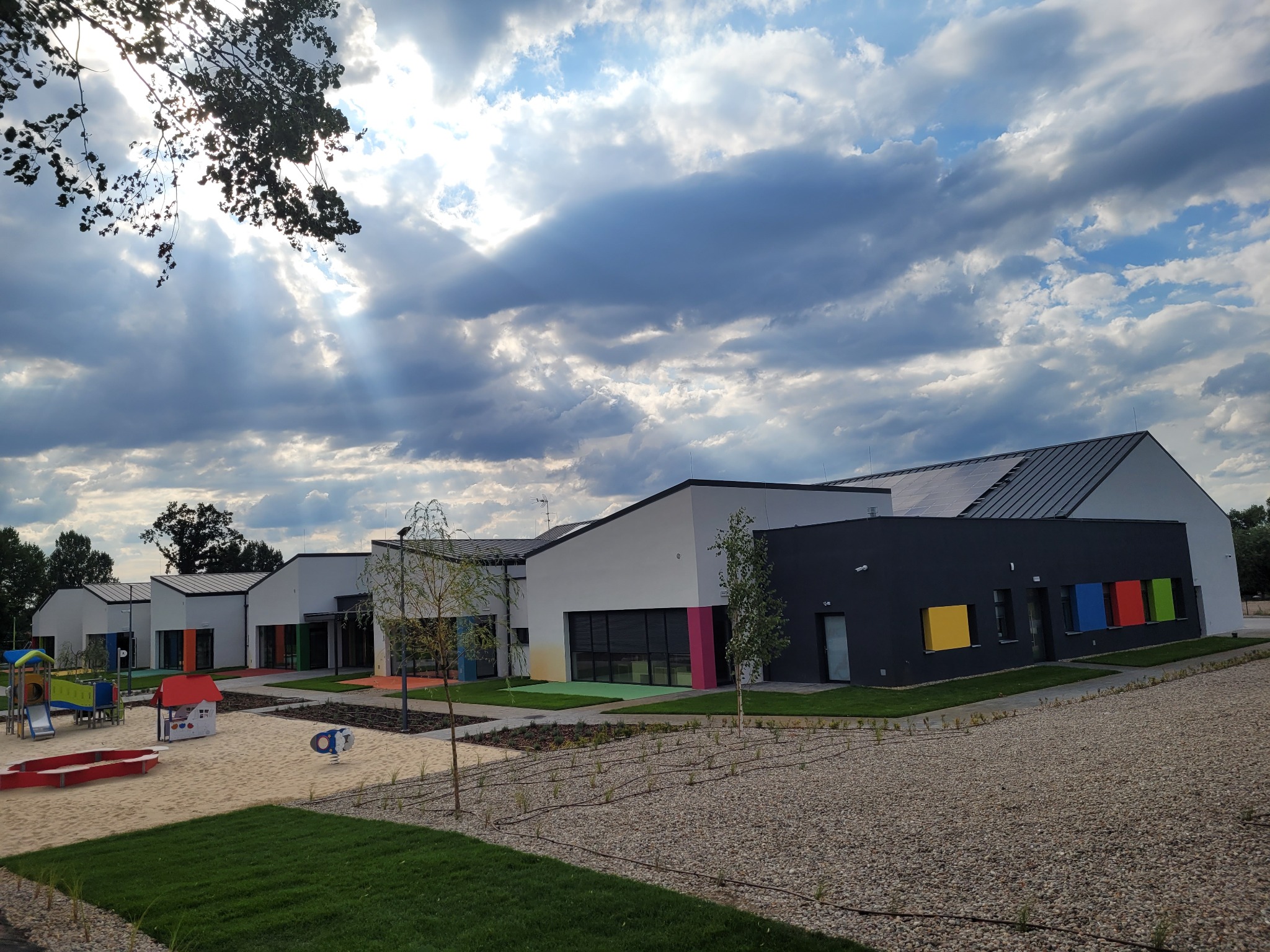 Nowoczesny budynek przedszkola z kolorowymi akcentami elewacji, placem zabaw i piaskownicą. Widok na fasadę w słoneczny dzień.