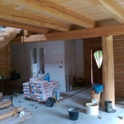 RAFFENER Rafał Jasiukajć - Wnętrze domu z bali w trakcie remontu, widoczne drewniane schody, sufit z belek z dekoracyjnymi wyżłobieniami i sterty worków z zaprawą na podłodze.