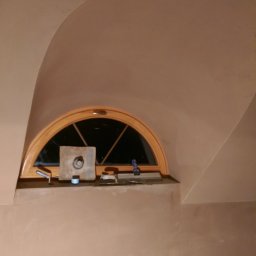 RAFFENER Rafał Jasiukajć - Wnętrze ze sklepieniem łukowym i półokrągłym oknem, ściany pokryte świeżo nałożoną, jasną gładzią, na parapecie okiennym narzędzia szpachlarskie i puszka z masą.
