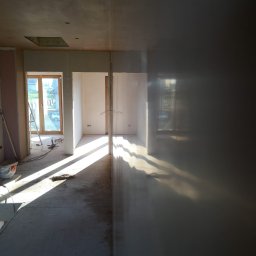 RAFFENER Rafał Jasiukajć - Wnętrze w trakcie prac wykończeniowych: surowe ściany, widoczne płyty kartonowo-gipsowe, szpachlowane powierzchnie, wpadające promienie słoneczne przez okno, drabina oparta o ścianę.