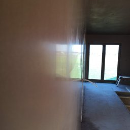 RAFFENER Rafał Jasiukajć - Świeżo wyszpachlowana ściana w trakcie prac wykończeniowych, widoczne okno z odbijającym się krajobrazem, szpachla z delikatnym połyskiem, przewody elektryczne wystające ze ściany, drabina w tle.