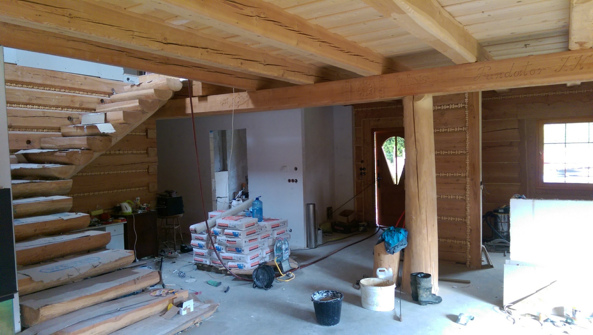 Wnętrze domu z bali w trakcie remontu, widoczne drewniane schody, sufit z belek z dekoracyjnymi wyżłobieniami i sterty worków z zaprawą na podłodze.