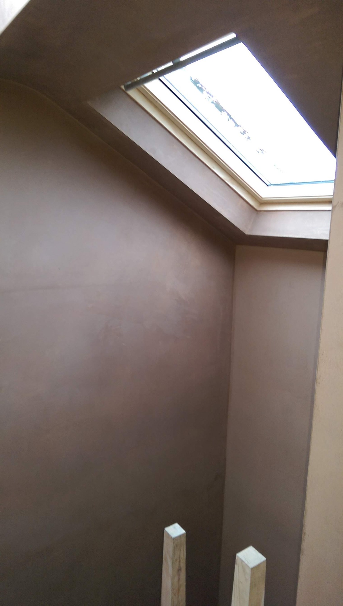 Świeżo nałożona, gładka, brązowa warstwa tynku na ścianie w pomieszczeniu z oknem dachowym, widoczne drewniane podpory u dołu kadru.