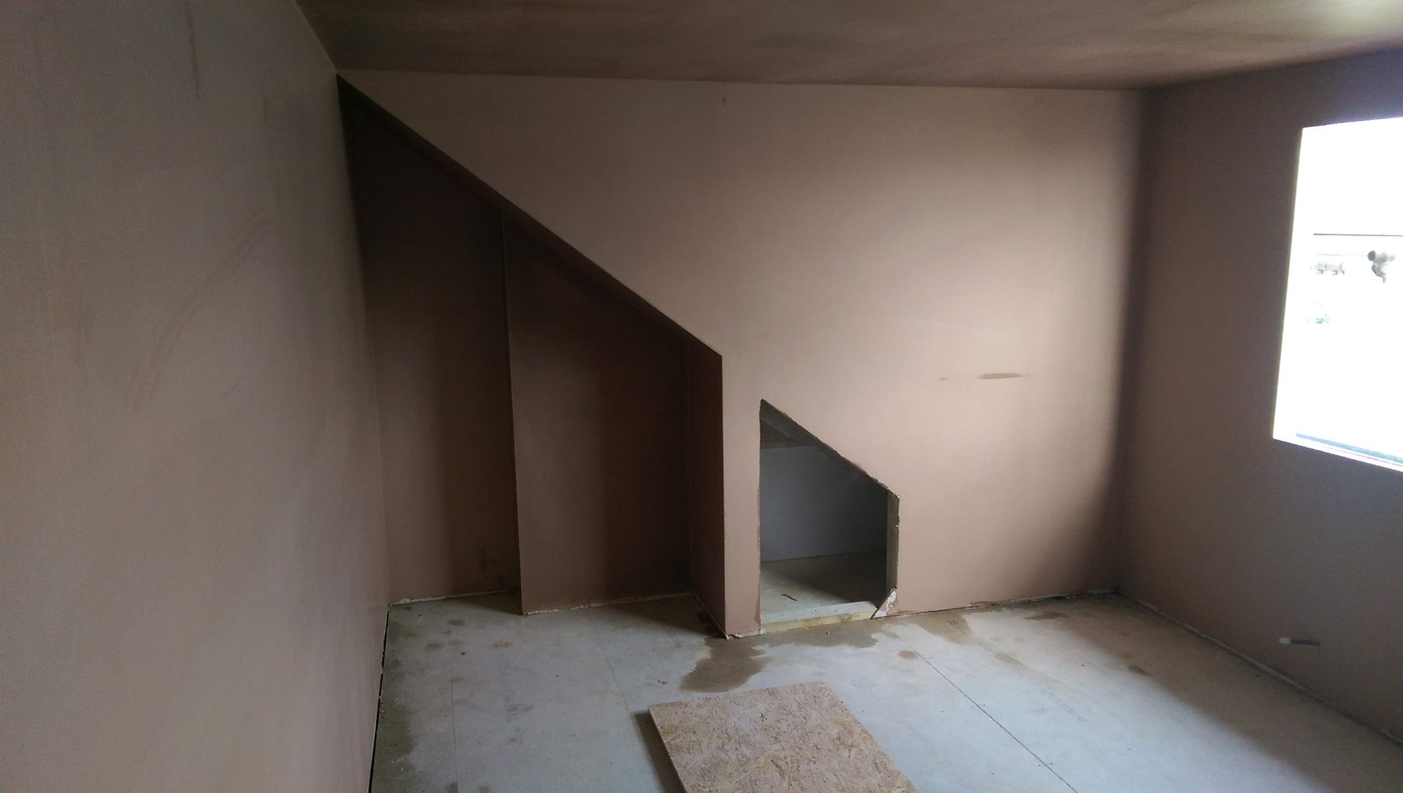 Pokój w trakcie prac wykończeniowych, ściany pokryte świeżo nałożoną, jeszcze nie wyschniętą gładzią, widoczny otwór w ścianie pod skosem.