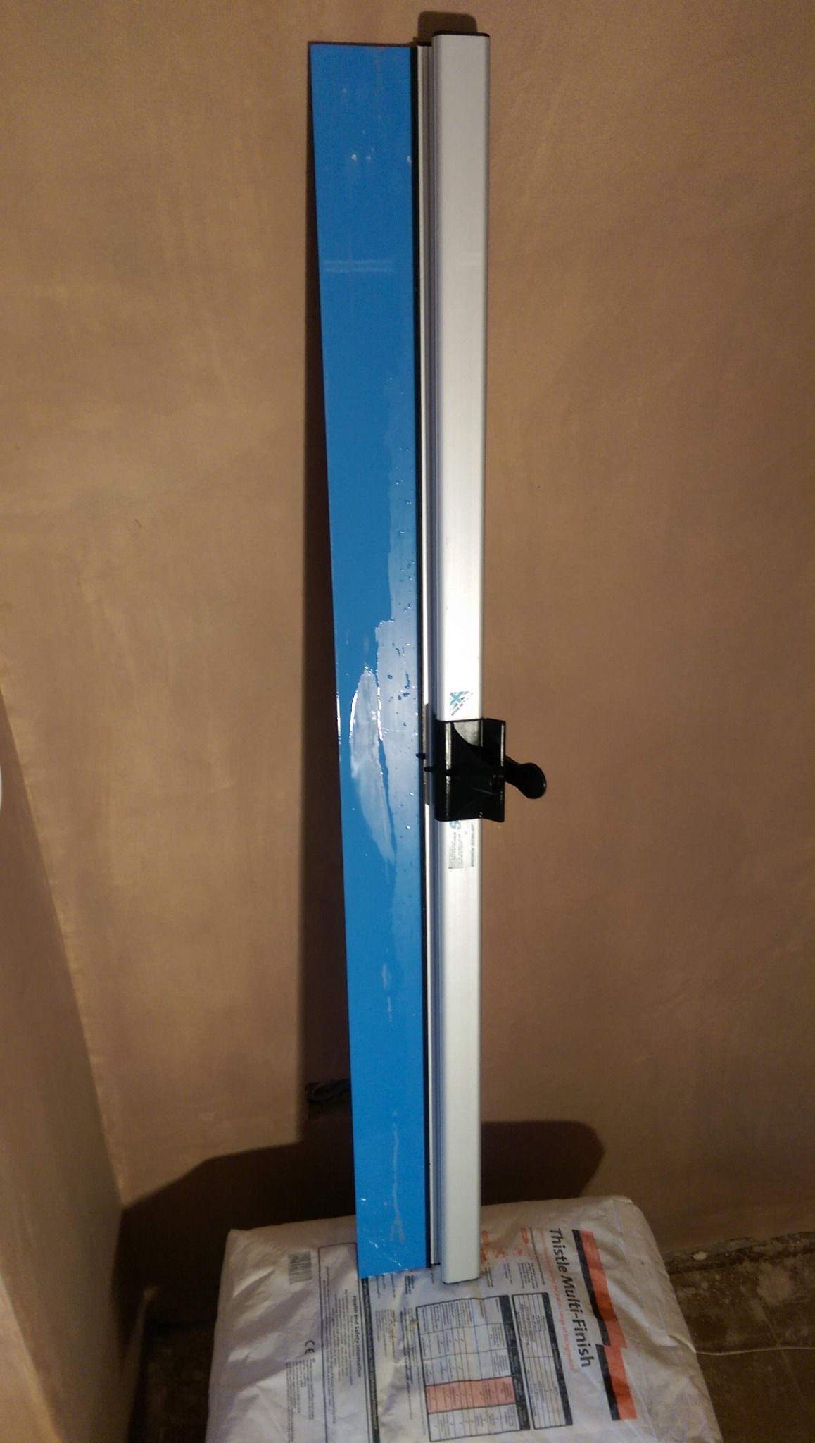 Aluminiowa listwa przypodłogowa z niebieskim elementem mocowana do ściany pokrytej świeżo nałożoną gładzią, stojąca na worku z gładzią szpachlową.